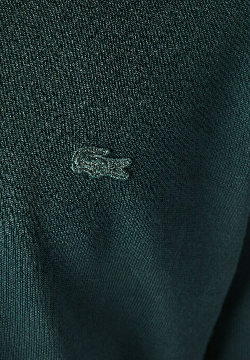 Tissu tricoté vert foncé avec une finition texturée, comportant un logo brodé ressemblant à un crocodile sur le côté gauche.