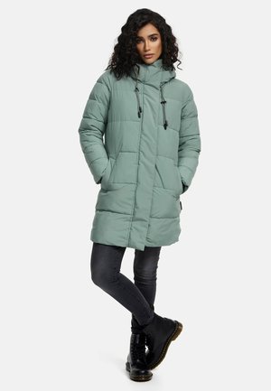 Manteau rembourré vert clair avec un col haut, des cordons de serrage réglables et des poches cachées. Associé à des bottes noires et un jean foncé.