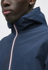 Ανεμοφρακτικό μπουφάν ναυτικού μπλε με κουκούλα, φερμουάρ μπροστά και πολύχρωμες ρίγες. Λείο, ελαφρύ υλικό με μοντέρνο σχέδιο.