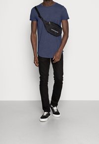 Svart axelväska med dragkedja och logotyp, bärs över en marinblå t-shirt. Slimmade svarta jeans och svarta skor med vita sulor fulländar looken.