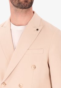 Giacca beige doppiopetto con revers a punta, due bottoni frontali e una tasca sul petto, realizzata in tessuto liscio.