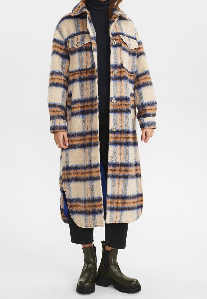 Frau in einem langen beigen Mantel mit blau-braunem Karomuster, schwarzen cropped Hosen, schwarzem Rollkragenpullover und dunkelgrünen Stiefeletten.