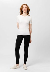 Bílý krátký rukáv těsně přiléhající top v kombinaci s černými legínami, obojí z hladkého materiálu. Model má na sobě světlé sportovní boty. Jednoduchý design.