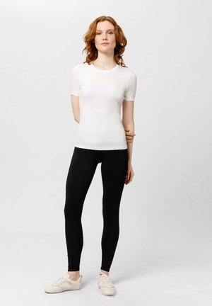 Haut ajusté à manches courtes blanc associé à un legging noir, tous deux en tissu lisse. Le modèle porte des chaussures de sport de couleur claire. Design simple.