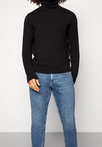 Suéter negro de cuello alto, hecho de una tela elástica, con un diseño ajustado, mangas largas y puños acanalados. Llevado con jeans azul claro.