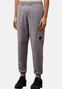 Helle graue Jogginghosen mit elastischem Bund, schmal zulaufenden Beinen, seitlicher Tasche mit einem schwarzen Knopf und glatter Baumwolloberfläche. Schwarze Schuhe sind sichtbar.