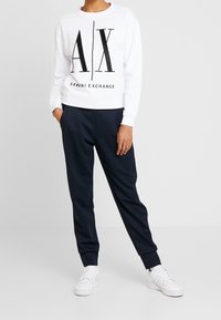 Felpa bianca con grafica nera "AX" e testo "ARMANI EXCHANGE", abbinata a pantaloni jogger blu navy, indossata con sneakers bianche.