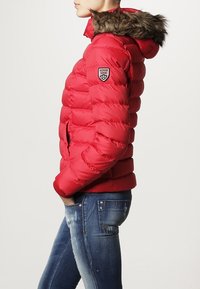 Rød pufferjakke med hætte foret med pels, kviltet tekstur og et logo-patch på ærmet. Har et tætsiddende design og ribbede ærmer.