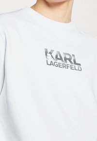 Weißes Baumwoll-T-Shirt mit einem metallisch silbernen Logo, das "KARL LAGERFELD" quer über die Brust liest. Klassisches Rundhals-Design, kurze Ärmel.