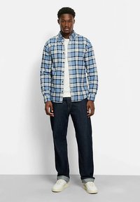 Karierter Hemd in Blau, Weiß und Beige, geknöpft, mit einem Kragen über einem weißen T-Shirt. Getragen mit dunklen Jeans und hellen Turnschuhen.