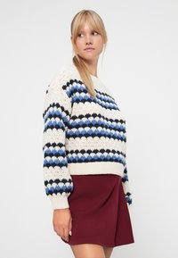Gestrickter Pullover in Creme mit blauem und schwarzem Zickzackmuster. Verfügt über lange, gepuffte Ärmel und einen gerippten Saum. Kombiniert mit einem burgunderfarbenen Rock.