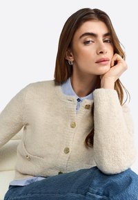 Cardigan beige realizzato in tessuto testurizzato, con scollo rotondo, bottoni dorati e due tasche frontali, indossato sopra una camicia a righe blu.
