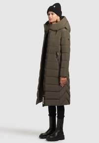 Vrouw in een olijfgroene gewatteerde lange jas met hoge kraag, zwarte beanie, zwarte legging en zwarte stevige laarzen, staande in de zij. 