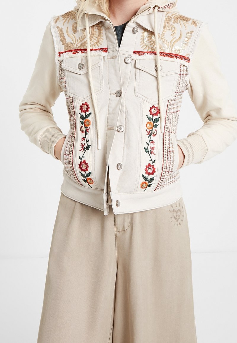 Femme portant une veste à capuche de couleur crème avec broderies florales et détails à motifs, associée à un pantalon beige ample.