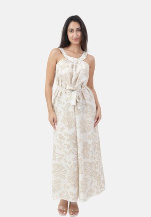 Femme portant une combinaison crème sans manches à jambes larges avec un imprimé floral beige et une ceinture nouée, posant les mains détendues sur les côtés.