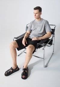 Svarte lærsandaler med kryssdesign, brukt sammen med grå t-skjorte og svarte shorts, sittende i en stol med kromramme på en lys bakgrunn.
