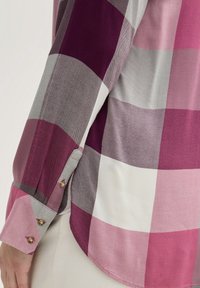 Nærbillede af en skjorteskjorte med lange ærmer med et stort ternet mønster i lyserød, hvid og lilla, samt to gyldne knapper på lyse beige bukser.