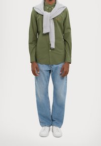 Man i olivgrön knapp-up skjorta med en grå tröja draperad över axlarna, ljusblå jeans och vita sneakers, stående.