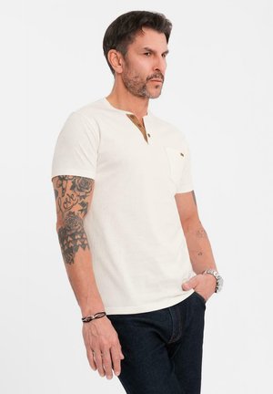 Camiseta blanca de manga corta hecha de algodón, con un pequeño bolsillo y botones de madera en el cuello. Llevada con jeans de mezclilla oscura.