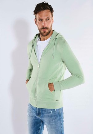 Light green zip-up hoodie with drawstring hood, two front pockets, and ribbed cuffs, worn over a white t-shirt and blue distressed jeans.