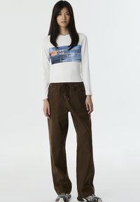 Witte langelijf top met een blauwe denim grafiek en een ronde halslijn, gecombineerd met losse bruine broek en zwarte sneakers.