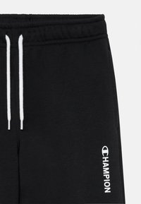 Svarte joggebukser laget av myk stoff, med snøring og hvite spisser, samt en hvit logo på nedre ben.