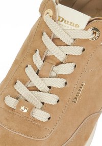 Beige suede sneaker with cream laces, featuring gold eyelets and logo accents. Smooth texture and rounded toe design.