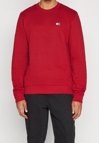 Sudadera roja con cuello redondo y puños y dobladillo acanalados, con un pequeño logo de Tommy Jeans en el pecho. Textura de tela suave.
