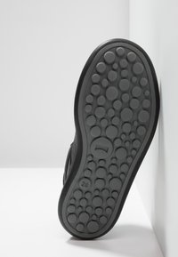 Suola di scarpa nera con superficie testurizzata che presenta motivi circolari. Numero '28' inciso vicino al tallone. Bordo liscio, inclinato contro un muro.