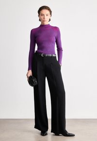 Camisola de malha canelada roxa com gola alta, combinada com calças pretas de perna larga e uma clutch preta. Botas de cano curto pretas com bico fino completam o visual.