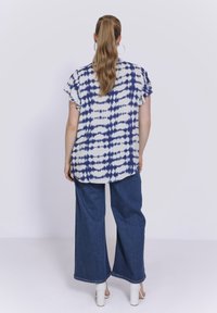 Kurzärmlige Bluse in Weiß und Marineblau mit Batikmuster, kombiniert mit weit geschnittenen blauen Jeans und weißen Blockabsatzsandalen. Glatter Stoff; lässige Passform.