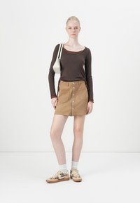 Braunes Langarmhemd, beiger Knopfrock mit Taschen, kombiniert mit gemusterten Schuhen und weißen Socken. Eine stylische Umhängetasche rundet das Outfit ab.