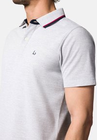 Szary polo z teksturowanej tkaniny, z granatowym kołnierzykiem ozdobionym czerwonymi i białymi akcentami. Małe logo wyszyte na klatce piersiowej.