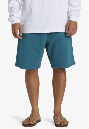 Shorts de sport teal avec une taille élastique et des poches latérales, offrant une texture lisse et une coupe décontractée, associés à des tongs.