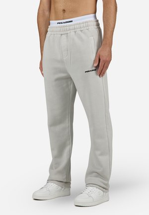 Pantalon de survêtement gris clair en matière douce, avec une taille élastique, des poches latérales et le logo noir "PEGADOR". Associé à des baskets blanches.