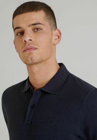 Polo shirt blu scuro con colletto a tre bottoni, tessuto testurizzato e logo sottile sul petto. Design aderente e maniche corte.
