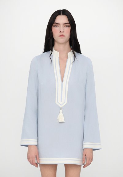 Robe tunique bleu clair avec un décolleté en V, des rayures blanches et un détail à pompon. Le tissu est lisse avec des manches longues et évasées.