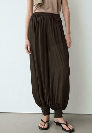 Personne portant un pantalon harem ample marron foncé avec des poignets froncés et des tongs noires, debout contre un mur blanc uni.