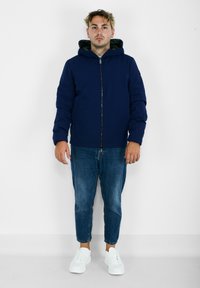 Giacca imbottita blu navy con zip frontale e cappuccio, abbinata a jeans azzurri chiari e sneakers bianche. Tessuto morbido e vestibilità comoda.