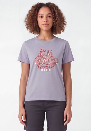 Frau mit lockigem Haar trägt ein lavendelfarbenes T-Shirt mit floralem Fahrrad-Design und dem Text "Life is a beautiful ride."