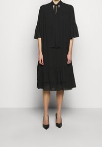 Robe noire à manches courtes avec col en keyhole et détail à nœud, présentant un ourlet à volants en couches et un tissu léger. Accompagnée de talons noirs pointus.