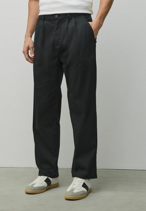 Man wearing black pleated pants, white t-shirt, and white sneakers with black stripes, standing against a light grey background.