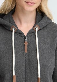 Sivá mikina na zips s kapucňou, bielymi šnúrkami a hnedými koženými detailmi. Obsahuje predný zips a mäkkú, textúrovanú látku.
