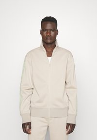 Felpa zip-up beige realizzata in tessuto morbido, con colletto alto e dettagli del logo verde sulle maniche. Silhouette aderente.