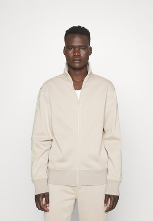 Sudadera con cremallera de color beige, hecha de un tejido suave, con un cuello alto y detalles de logo verde en las mangas. Silueta ajustada.