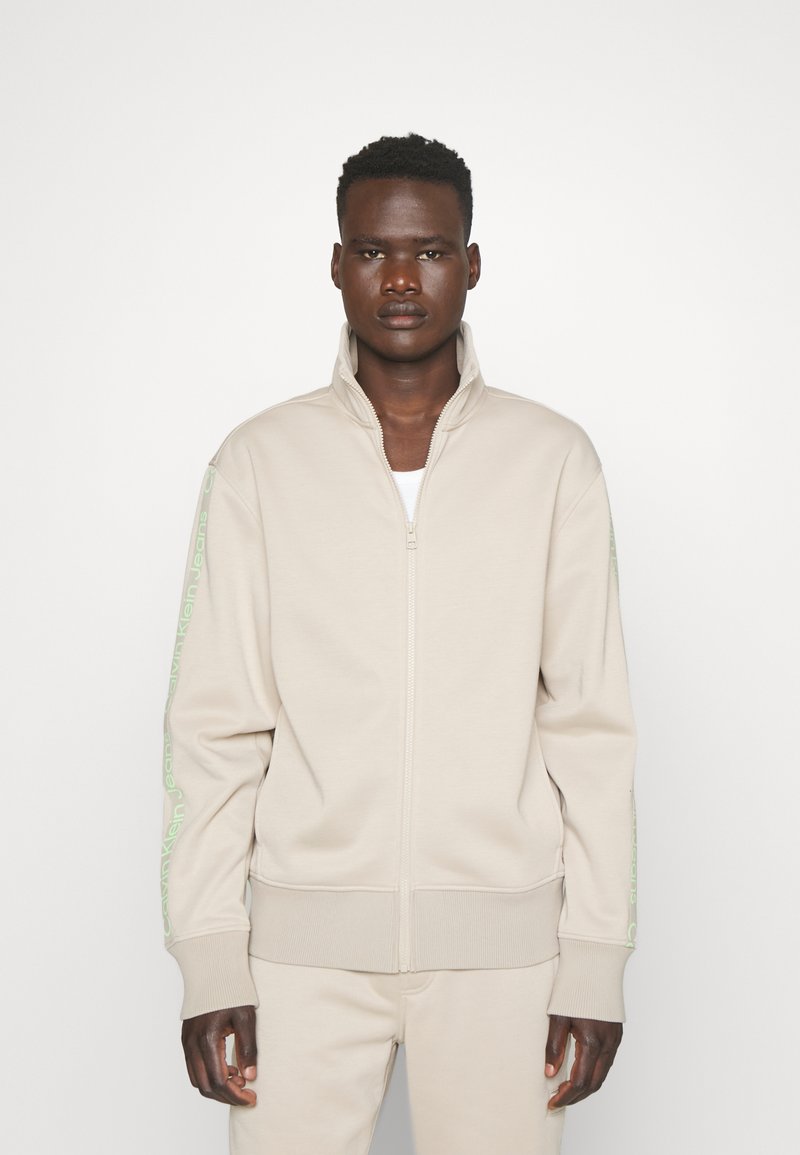 Felpa zip-up beige realizzata in tessuto morbido, con colletto alto e dettagli del logo verde sulle maniche. Silhouette aderente.