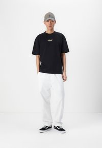 T-shirt in cotone nero con un piccolo logo giallo, abbinato a pantaloni bianchi a vestibilità ampia e sneakers nere Vans. Un cappellino da baseball grigio completa l'outfit.