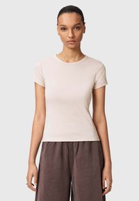 Jonge vrouw met haar in een staart draagt een nauwsluitend beige topje met korte mouwen en losse donkerbruine broek, tegen een effen achtergrond.