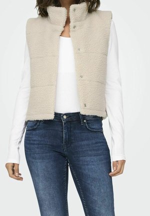 Femme portant un gilet en polaire sherpa beige sur un haut à manches longues blanc et un jean en denim bleu foncé, debout devant un fond uni.