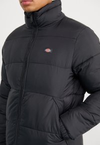 Giubbotto a vento nero con colletto alto, cerniera completa, motivo trapuntato orizzontale e patch con logo sul petto, caratterizzato da una texture liscia e leggera.